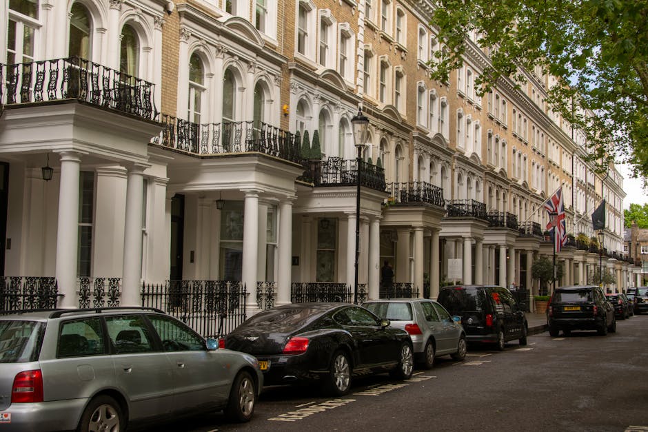 A row of elegant, white Victorian-style townhouses with black wrought iron balconies and columns along Brompton Road SW3. The ground floors feature large glass windows and entrances, while small potted plants and neatly maintained facades add charm. Parked cars, including a silver station wagon, a black sedan, and other vehicles, line the street in front of the buildings. The street is shaded by mature trees, with green leaves partially visible in the upper right corner. The setting appears quiet and well-kept, reflecting typical residential and commercial areas receiving regular surface cleaning and maintenance. The lighting is natural, suggesting daytime conditions. This scene is associated with professional cleaning services offered by Carpet Cleaners Brompton, focusing on surface cleaning and deep cleaning for flats and properties along Brompton Road.