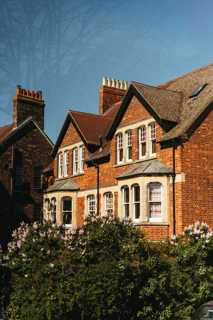 A row of traditional red brick residential houses featuring pitched tiled roofs, white-framed sash windows, and chimney stacks, set against a clear blue sky with some leafless trees in the background.